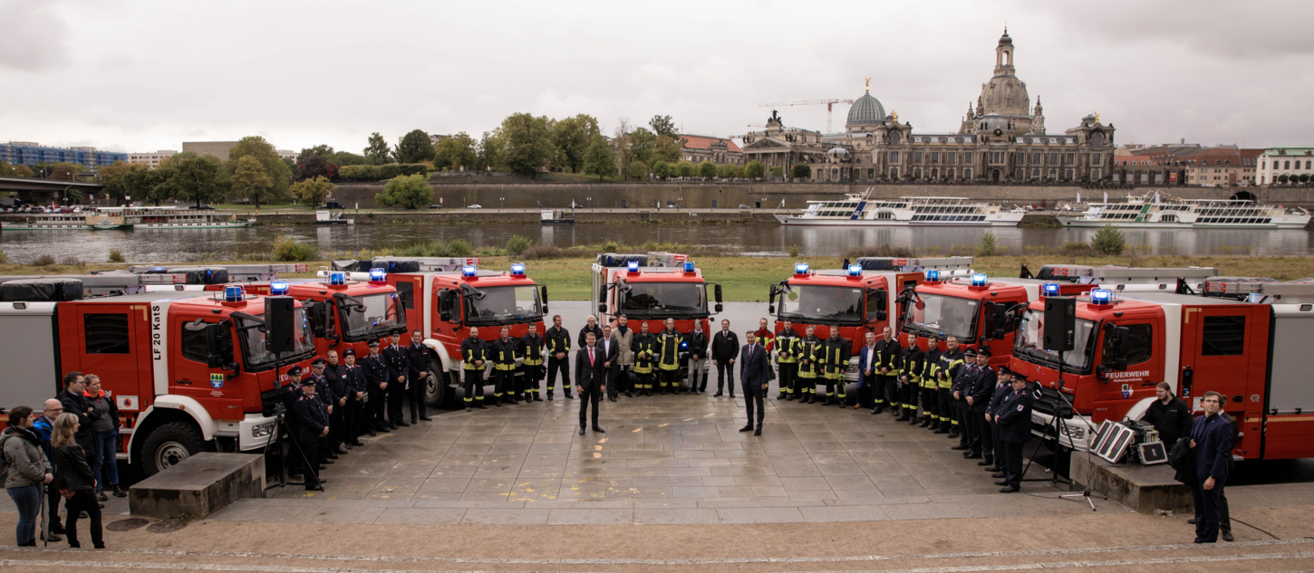 Sieben Katastrophenschutzfahrzeuge stehen in Form eines Halbkreises angeordnet vor der Dresdner Altstadt-Kulisse, davor alle Teilnehmer der Veranstaltung.